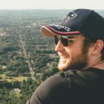 Smiling man in sunglasses and cap enjoying a scenic outdoor view from a hilltop.