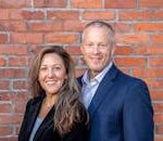 A cheerful middle-aged man and woman in business attire posing against a brick wall, depicting professionalism and partnership.