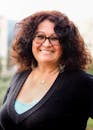 Portrait of a woman with curly hair and glasses smiling warmly in an outdoor setting.